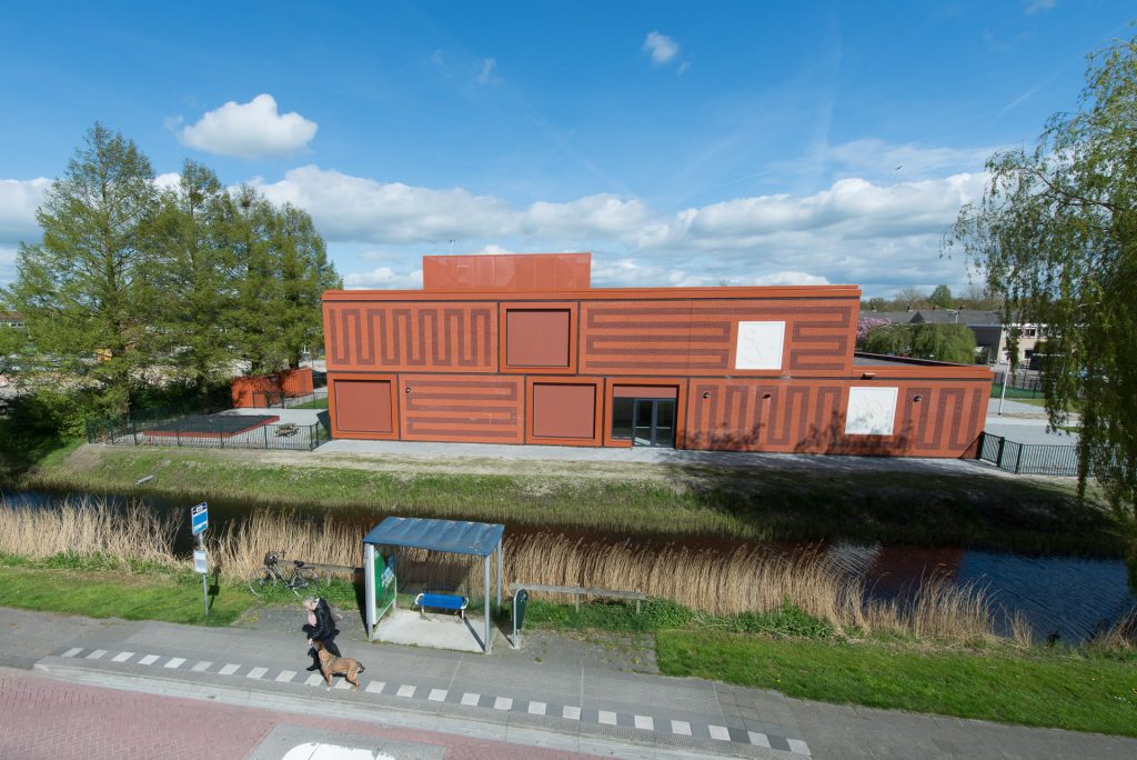 Rood gebouw met vlakken en tekening in het metselwerk aan een vaart met een bushalte op de voorgrond