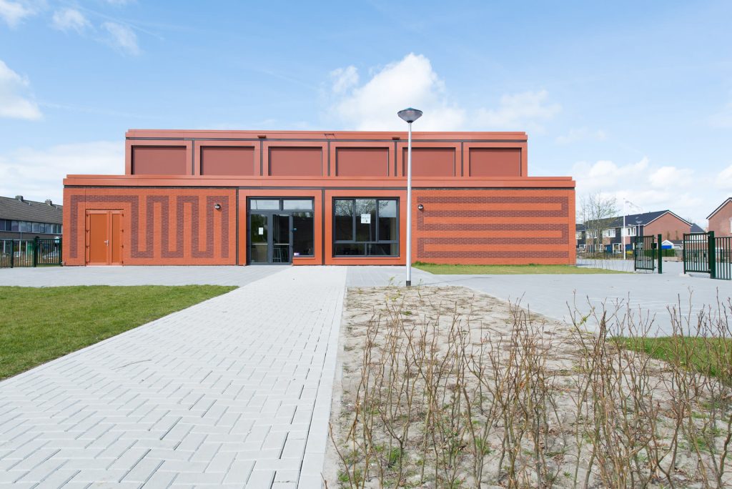 Vooraanzicht van rood gebouw met rode voeg en tekening in het metselwerk en plein op de voorgrond