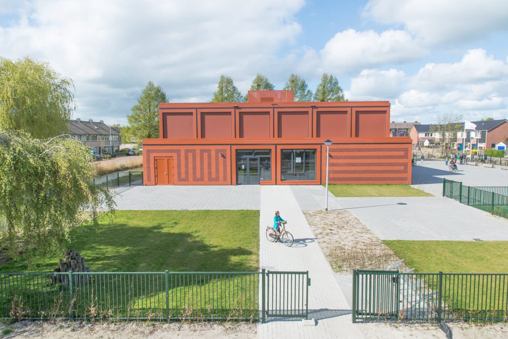 Vooraanzicht van rood gebouw met rode voeg en tekening in het metselwerk en plein op de voorgrond met fietser