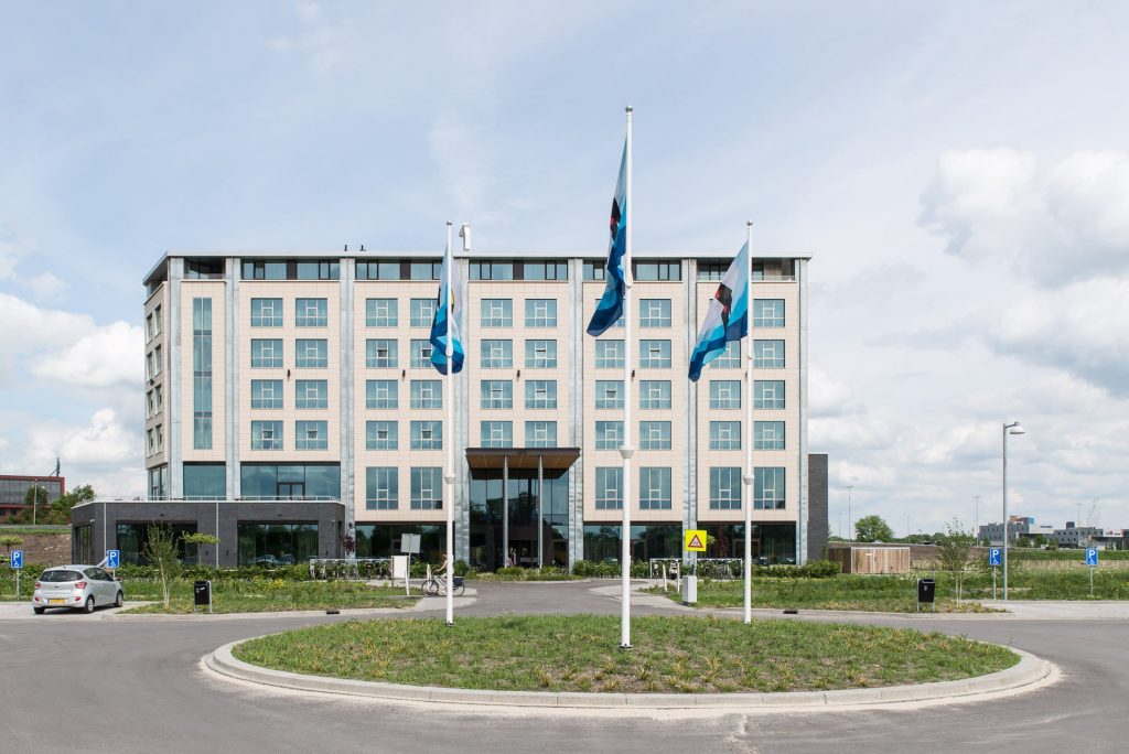Witte gevel van het Van der Valk hotel met een rotonde op de voorgrond waarin 3 vlaggenmasten staan.