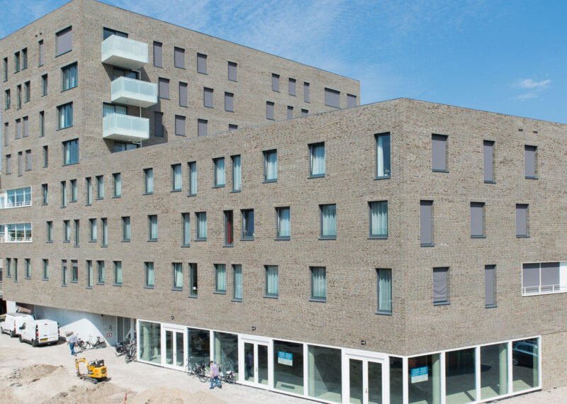 Zijaanzicht van hoogbouw in een bruingrijze steen met veel ramen tegen een blauwe lucht