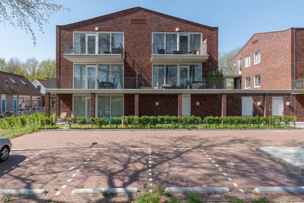 Rechte aanblik van een roodbruin gebouw met een puntdak, ramen en balkons. Op de voorgrond parkeerplaatsen.