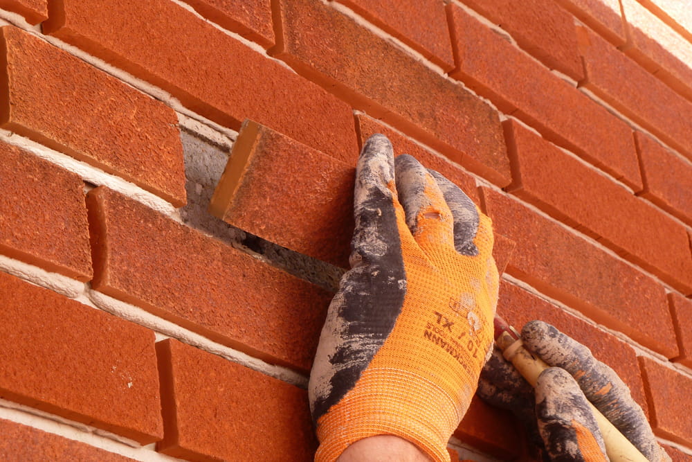 Handen met werkhandschoenen plakken een laatste steenstrip op een rode muur