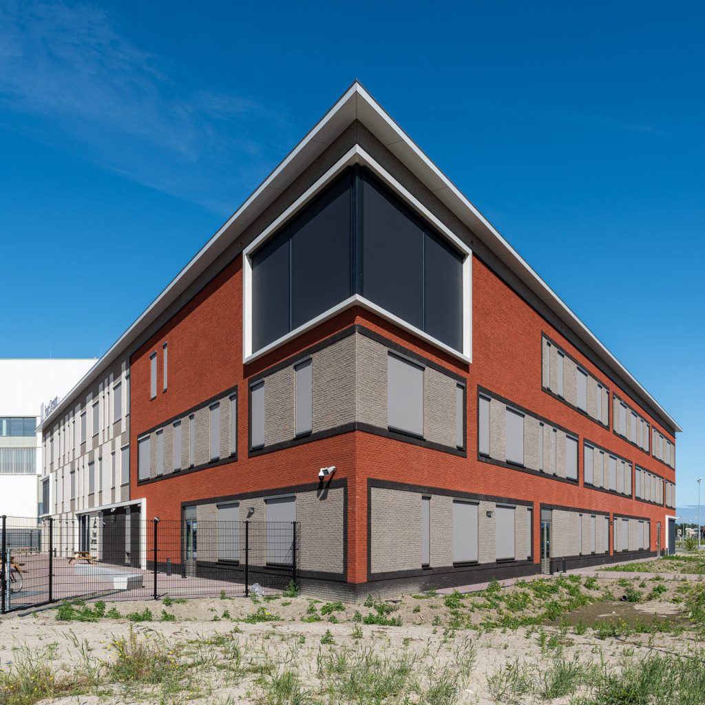 Hoek van een gebouw met rode en lichte vlakken, met groen en zand op de voorgrond tegen een blauwe lucht