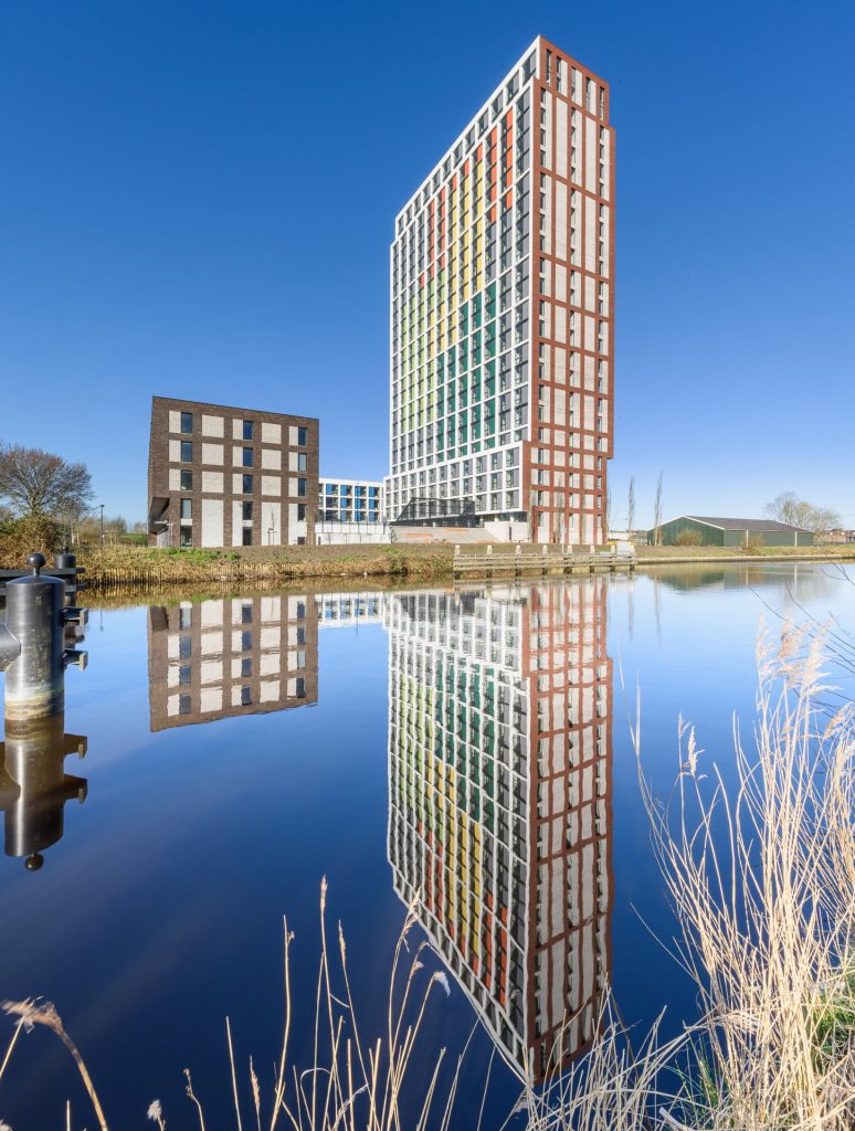 Een toren van rode en witte vlakken baksteen en gekleurde vlakken tegen een blauw lucht en op de voorgrond water